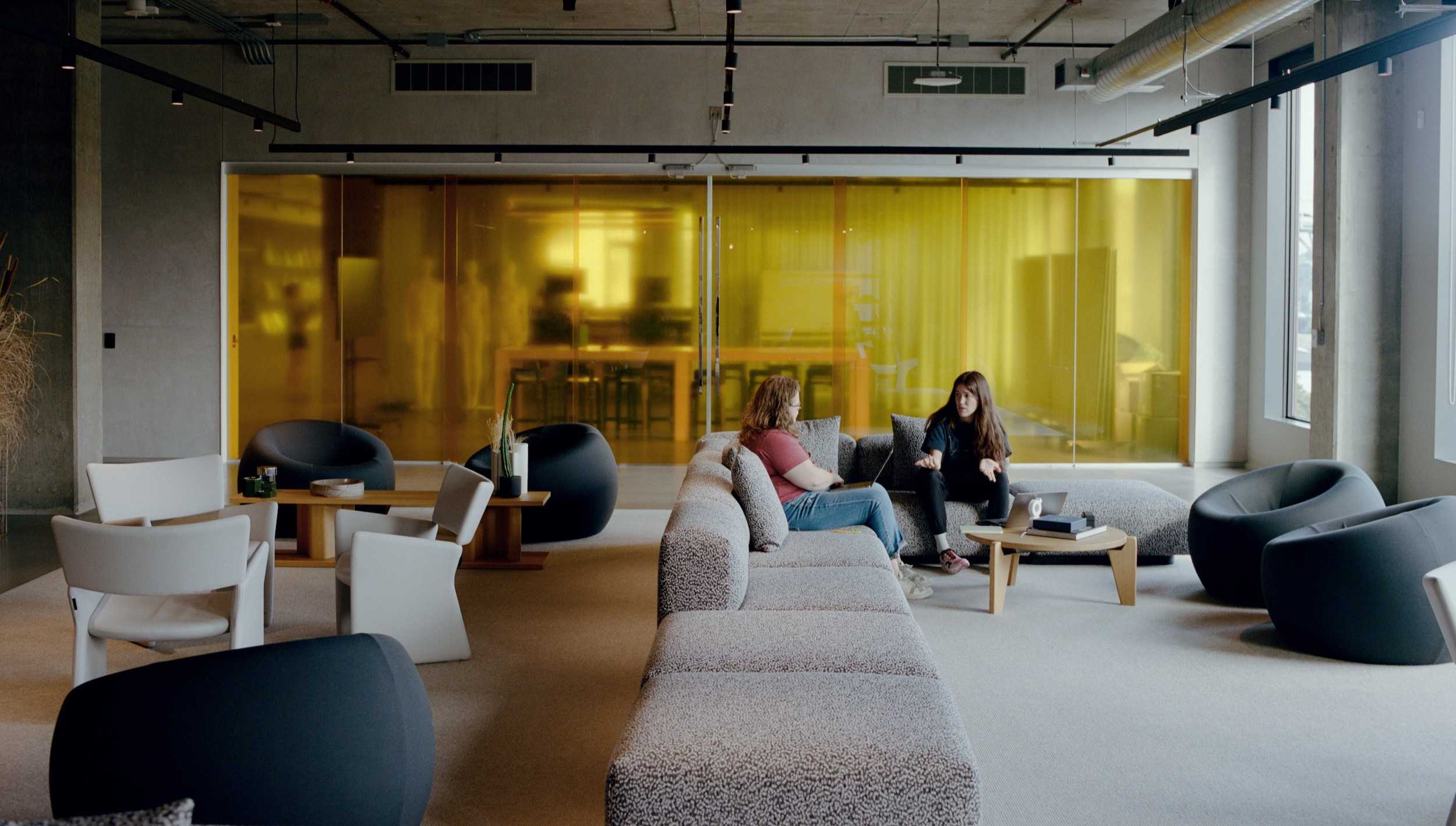 Two people seated on a long gray sectional sofa talking in a modern office lounge with a yellow-tinted glass wall and low tables.