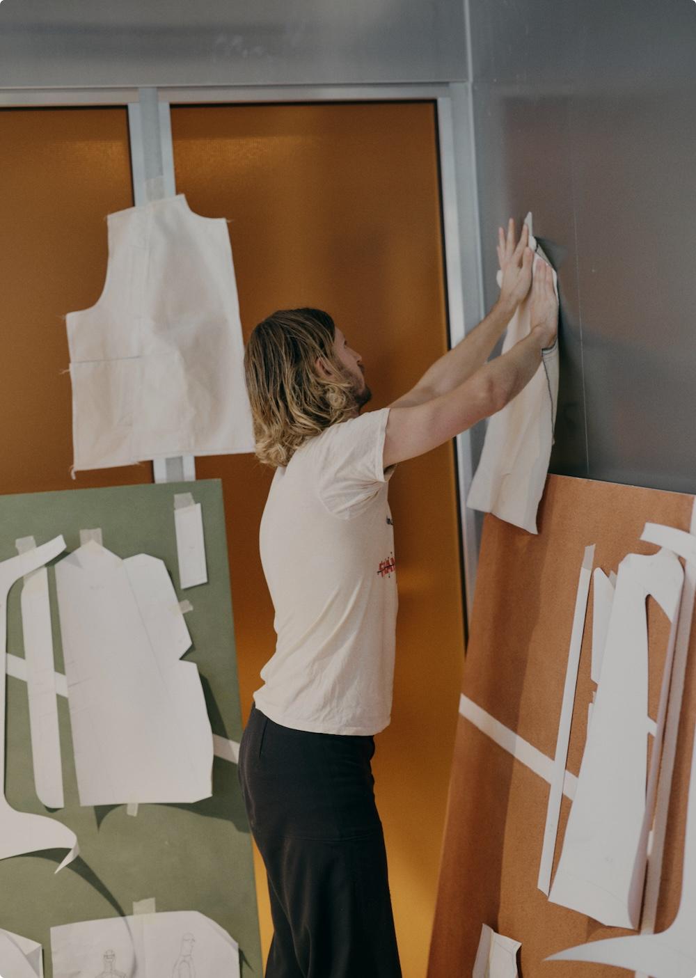 A person presses a fabric or paper pattern onto a wall while garment pattern pieces are taped to nearby boards.