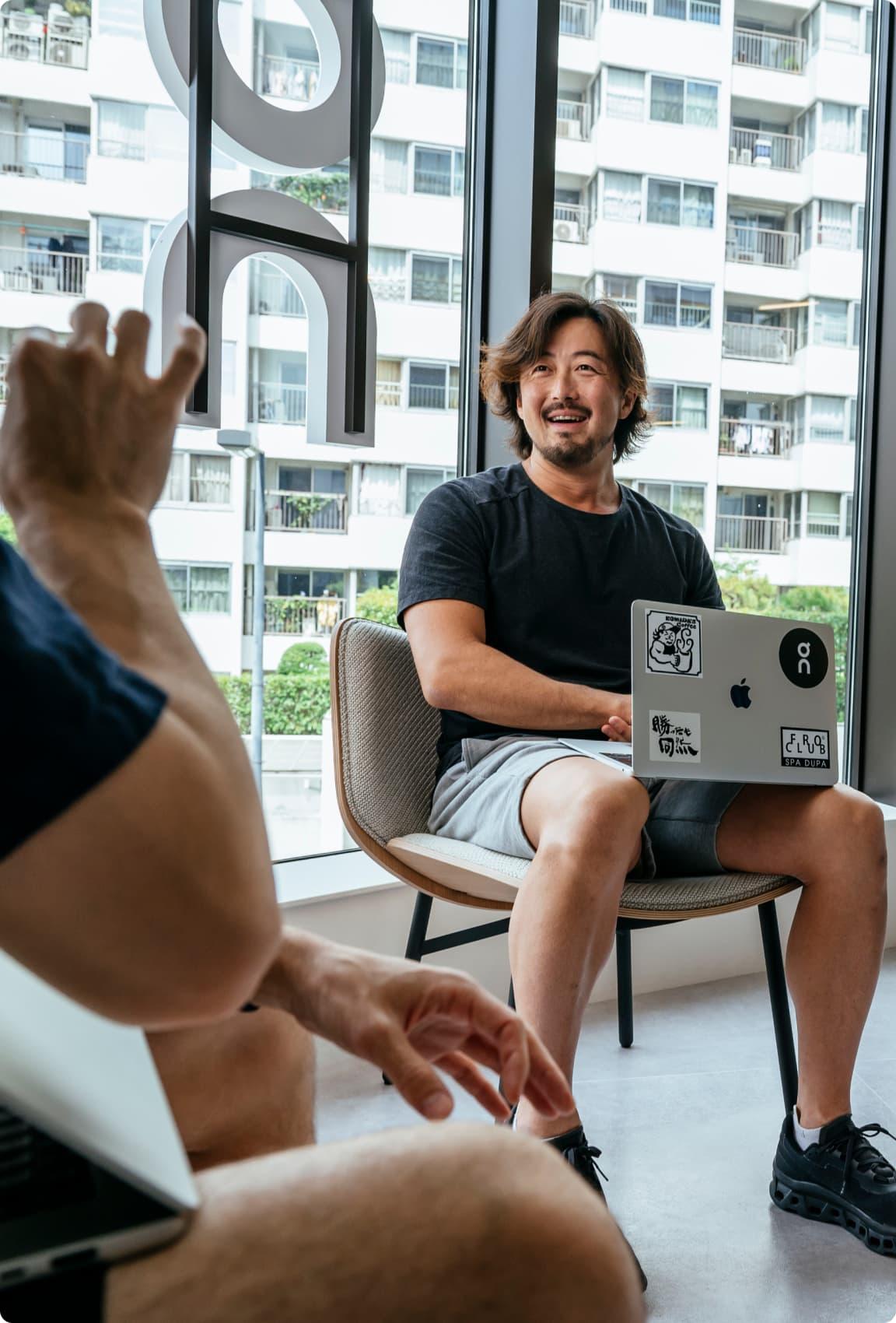 Man smiling while seated with a sticker-covered laptop on his lap, engaging in conversation with another person in a modern office with large windows and city apartment view.