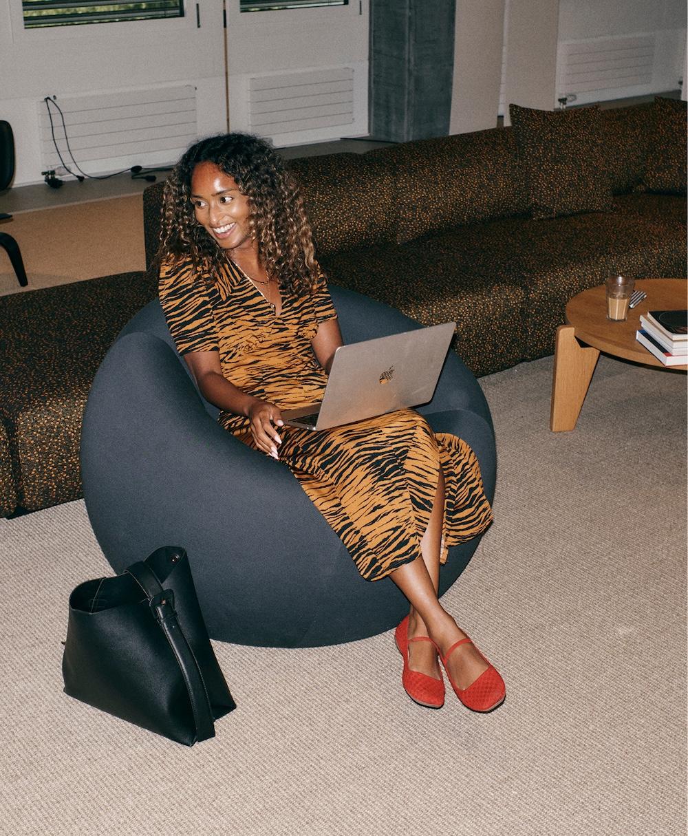 A smiling woman in a tiger-striped dress sits in a gray beanbag chair using a laptop, with a black tote on the floor and a coffee table with books and a drink beside a patterned sofa.