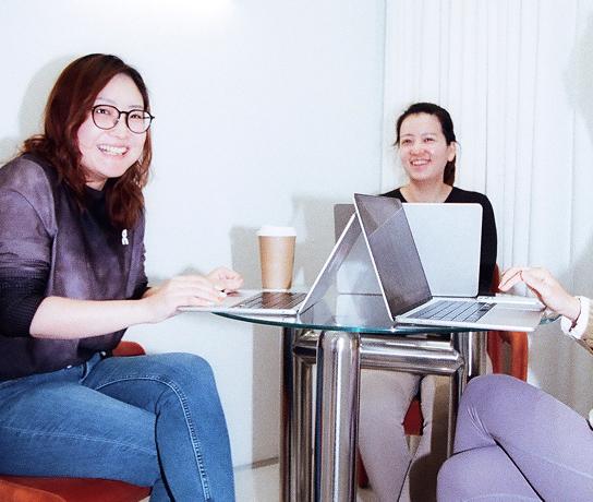 Three people sit around a glass table with open laptops and a paper coffee cup, smiling.