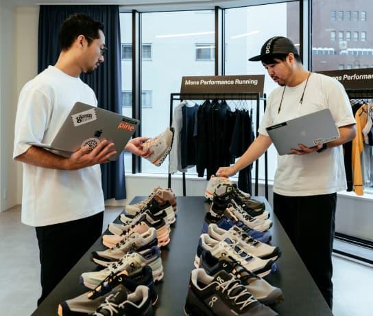 Two men with laptops in hand pick up and look at two rows of On Running shoes.