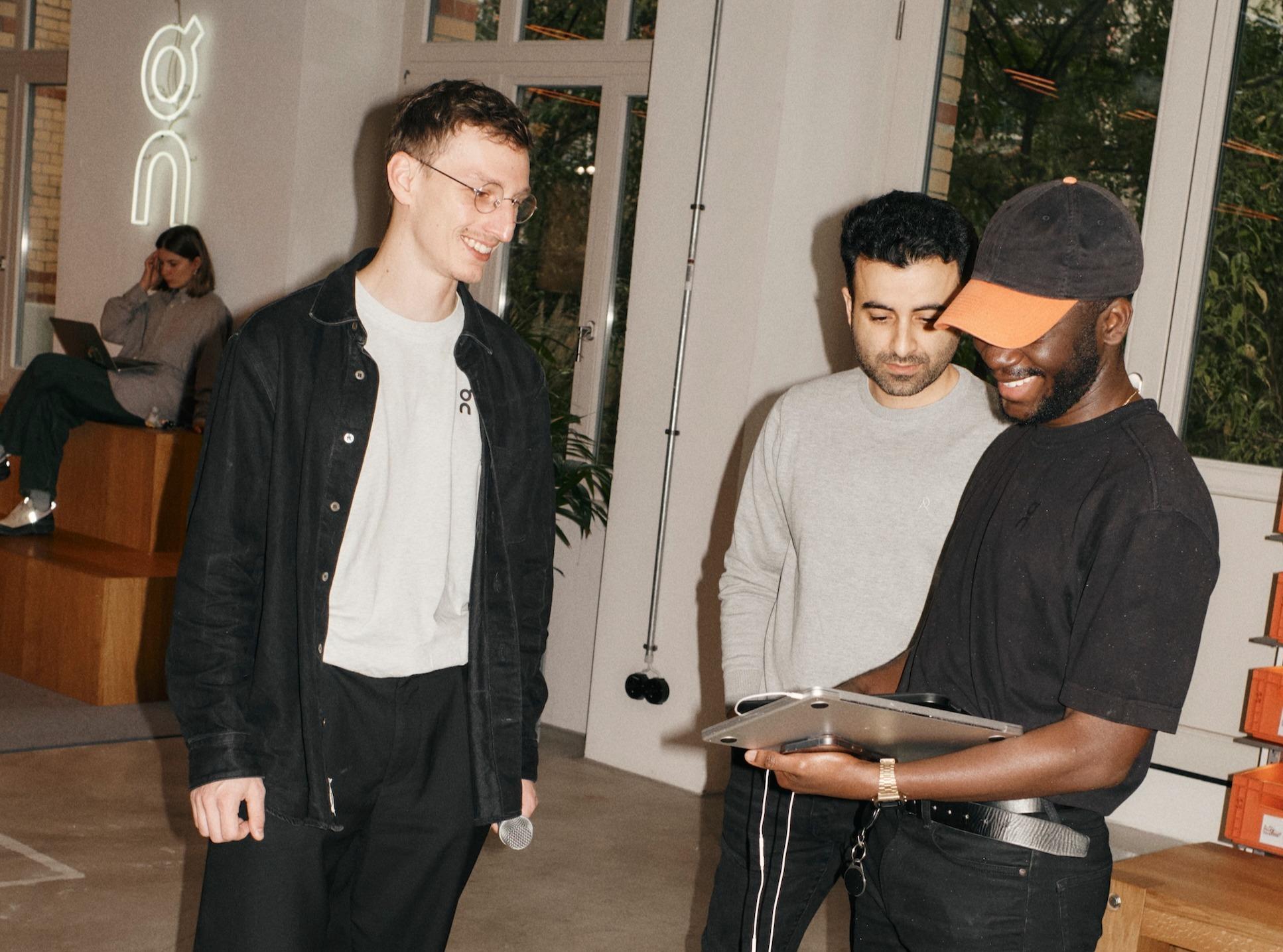 Three men stand together looking at a laptop one of them holds while a woman sits on wooden steps in the background using a laptop.