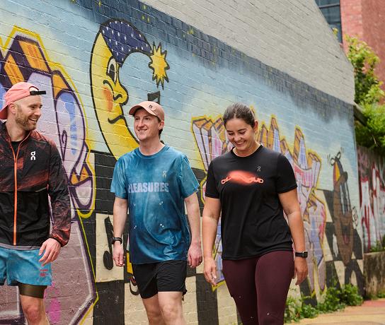 Three people in athletic clothing walk and smile past a colorful graffiti mural painted on a brick wall.