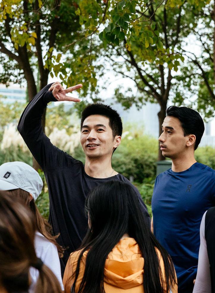 Man in a black long-sleeve shirt smiling and making a peace sign while standing in a group of people outdoors, with green trees and tall grass in the background.