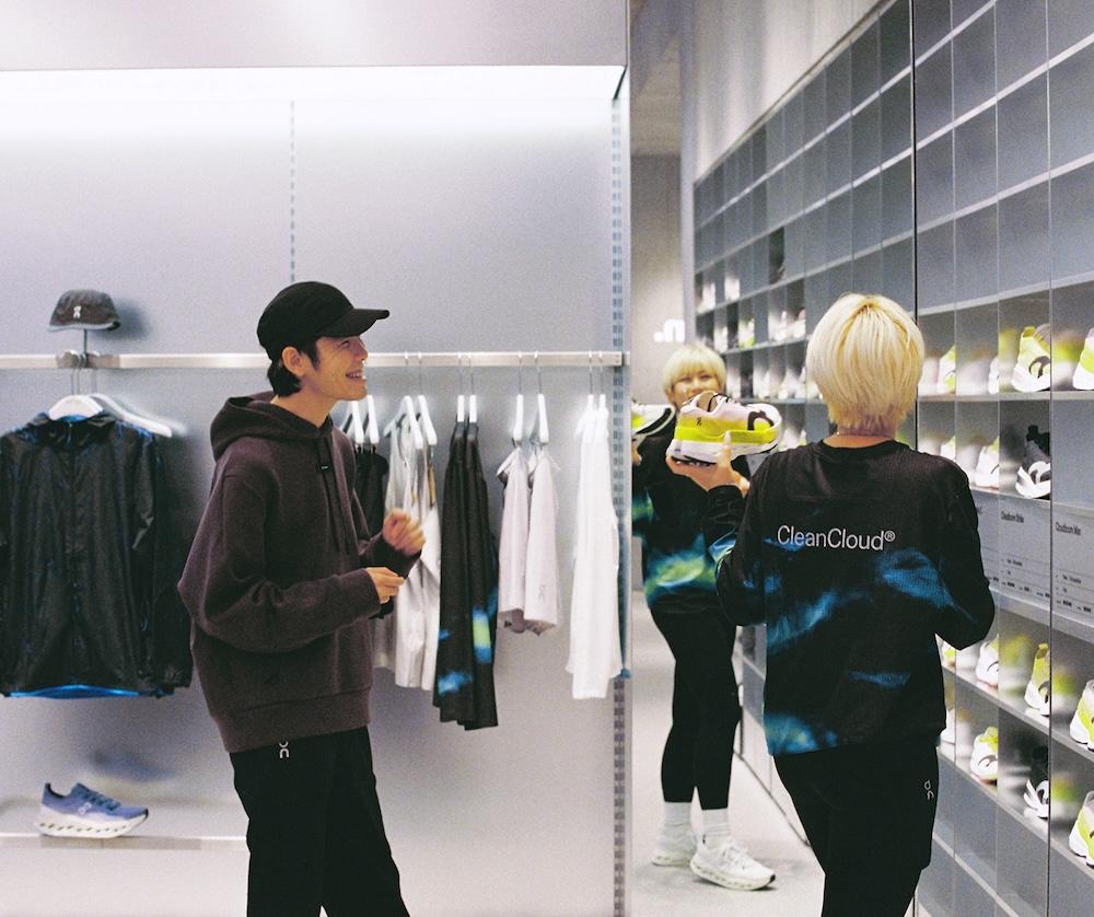 Two people in a retail store look at a neon yellow sneaker, with clothing on a rack to the left and shelves of shoes to the right.