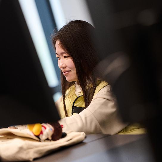 A person wearing a yellow vest and light-colored sweatshirt sits at a table, smiling as they look off to the side, with a cloth bag and a small stuffed toy in the foreground.