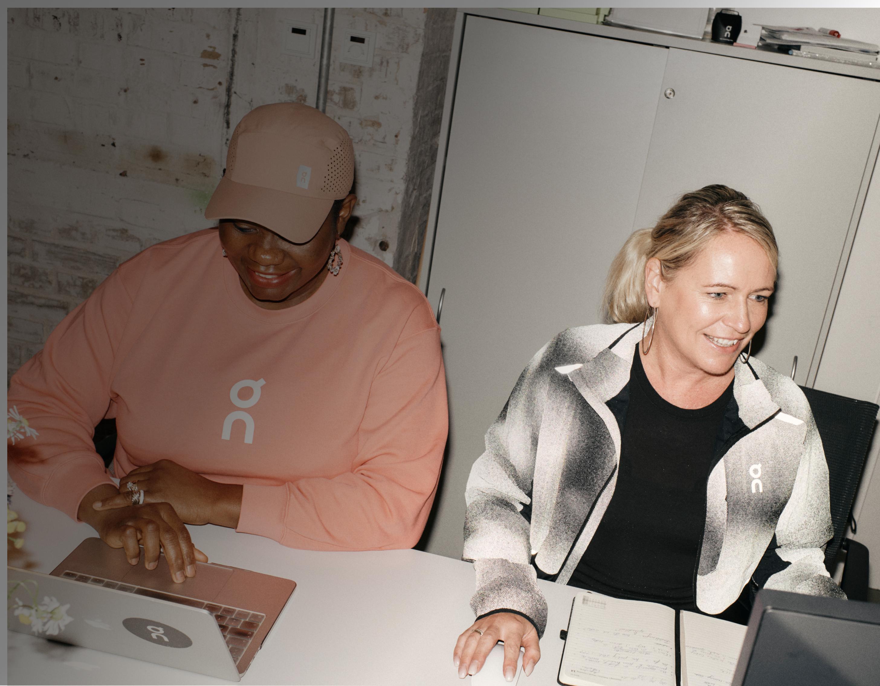 Two women smile while working at laptops in a casual office, both wearing On-branded apparel.