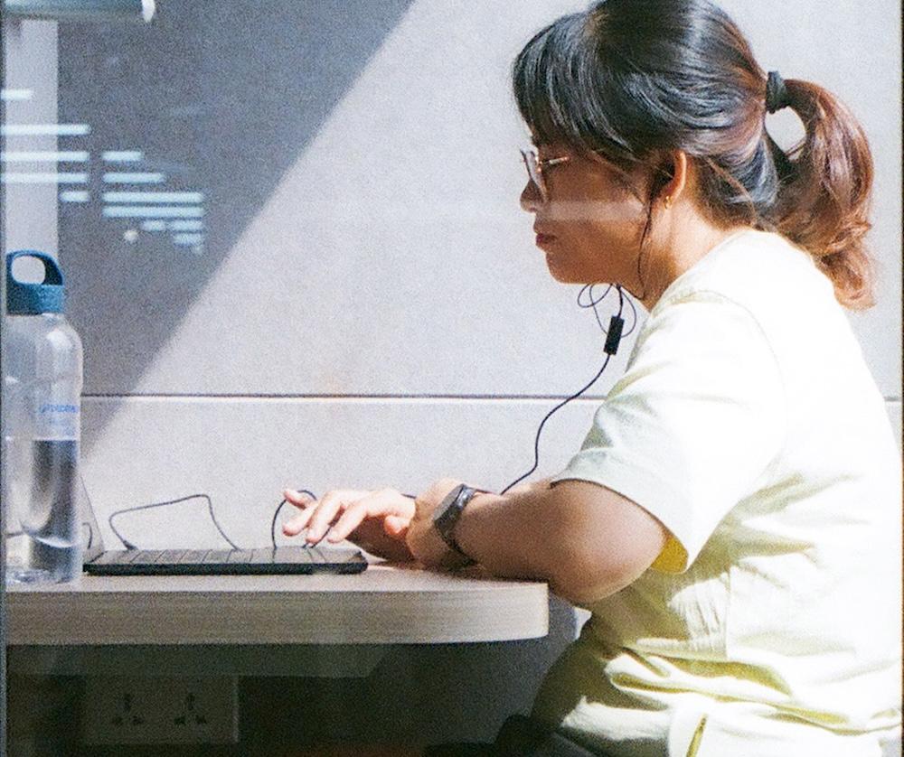 A person with glasses and a ponytail sits at a table typing on a laptop while wearing earphones, with a water bottle on the table.