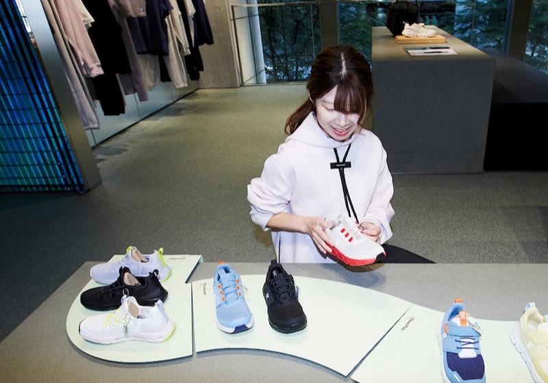 Smiling woman in a light hoodie holds a red and white sneaker while browsing colorful shoes displayed on a curved table in a store.