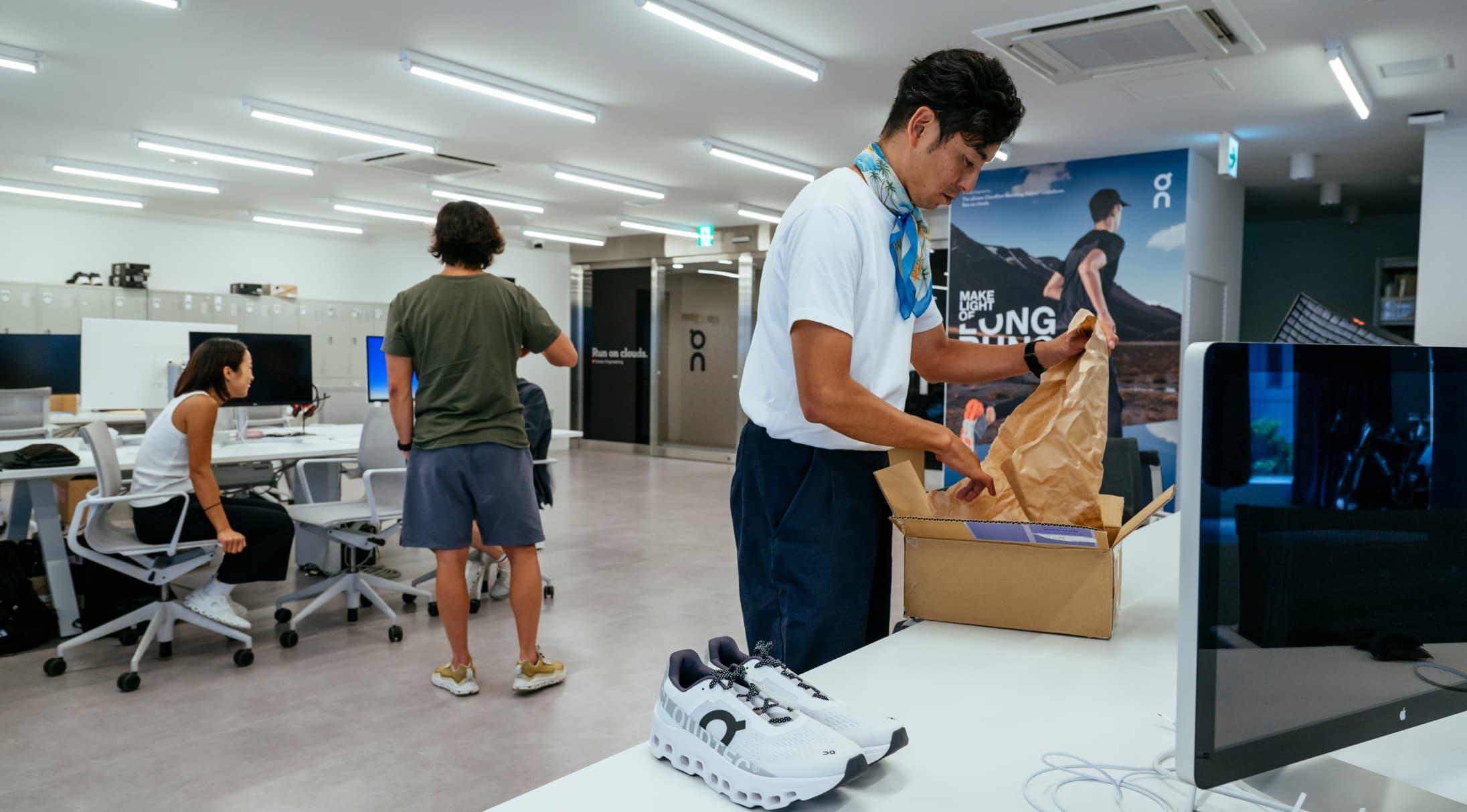 A man unpacks a box with an On Running shoe on the desk next to him.
