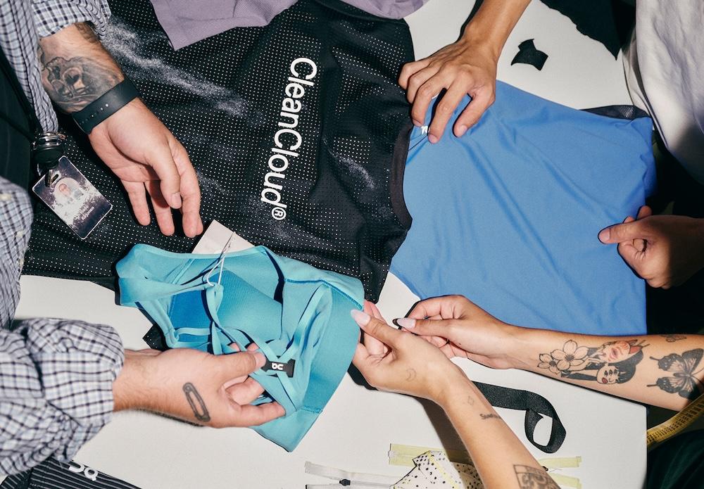 Top-down view of multiple hands examining blue clothing pieces next to a black mesh bag labeled "CleanCloud" on a table.