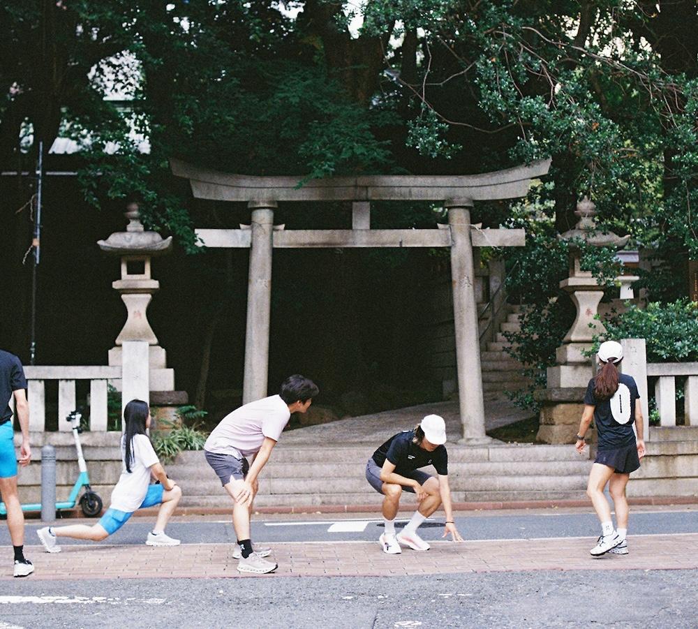 People in athletic clothing stretch on a sidewalk in front of a stone torii gate flanked by stone lanterns.