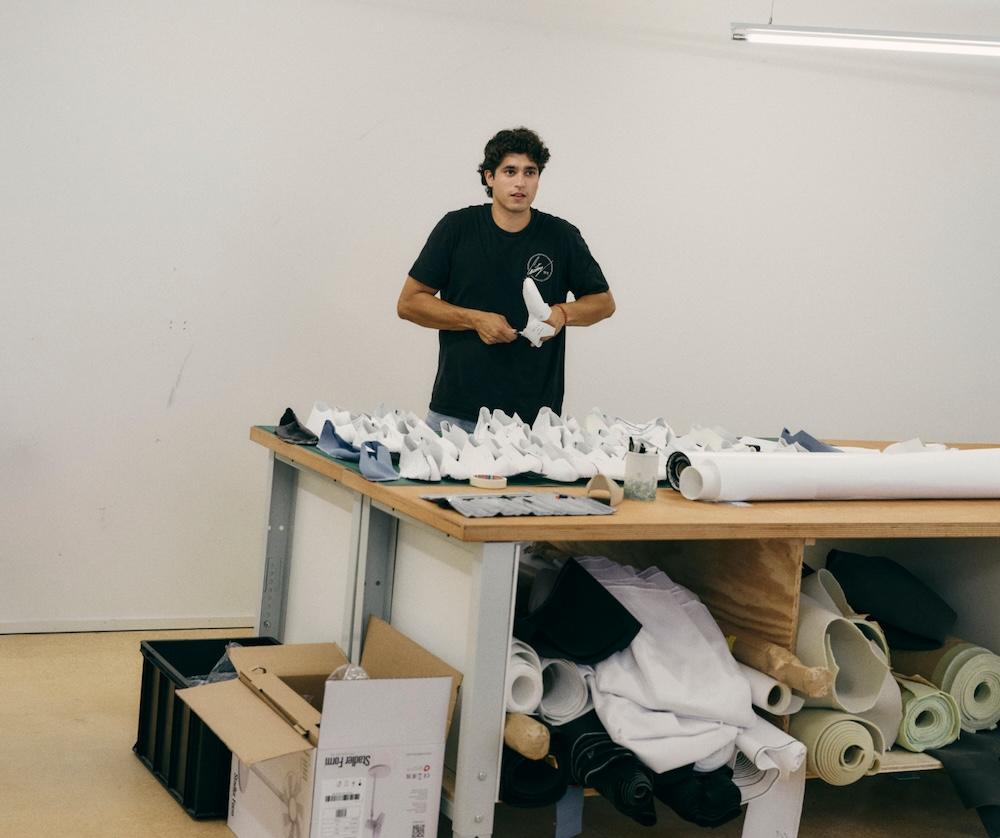 A man stands behind a large workbench arranging rows of white shoe forms, with rolls of fabric and cardboard boxes stored underneath and nearby.