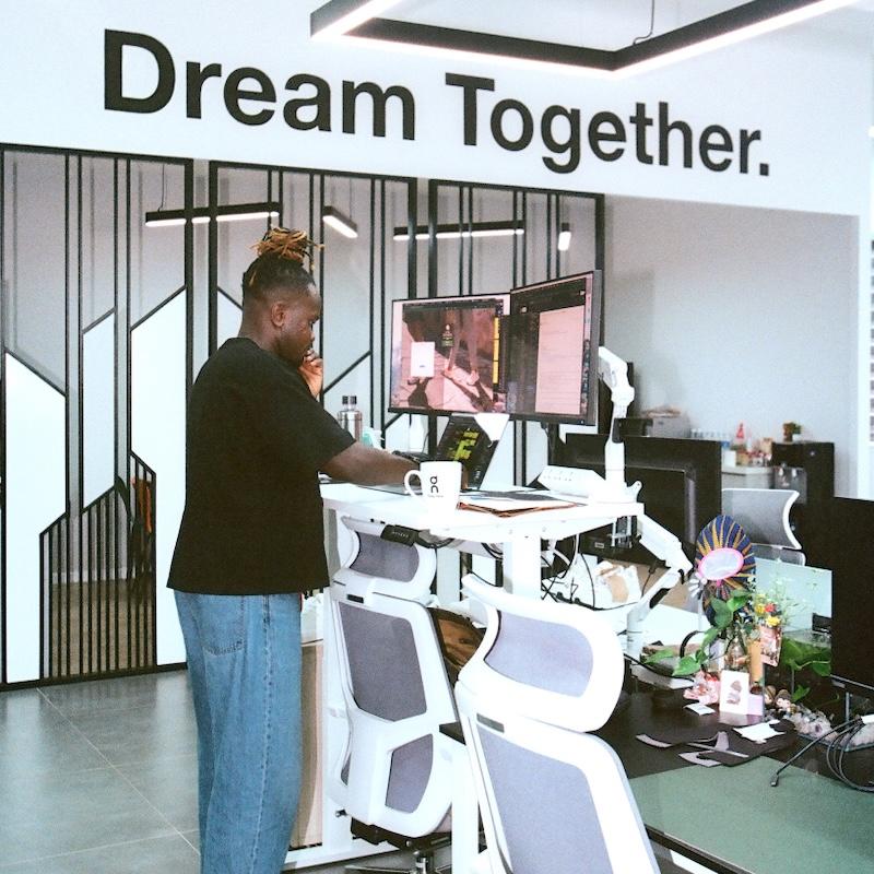 A person stands at a white adjustable standing desk with two monitors, white office chairs, and assorted desk items in an open office beneath a wall sign reading 'am Together.'