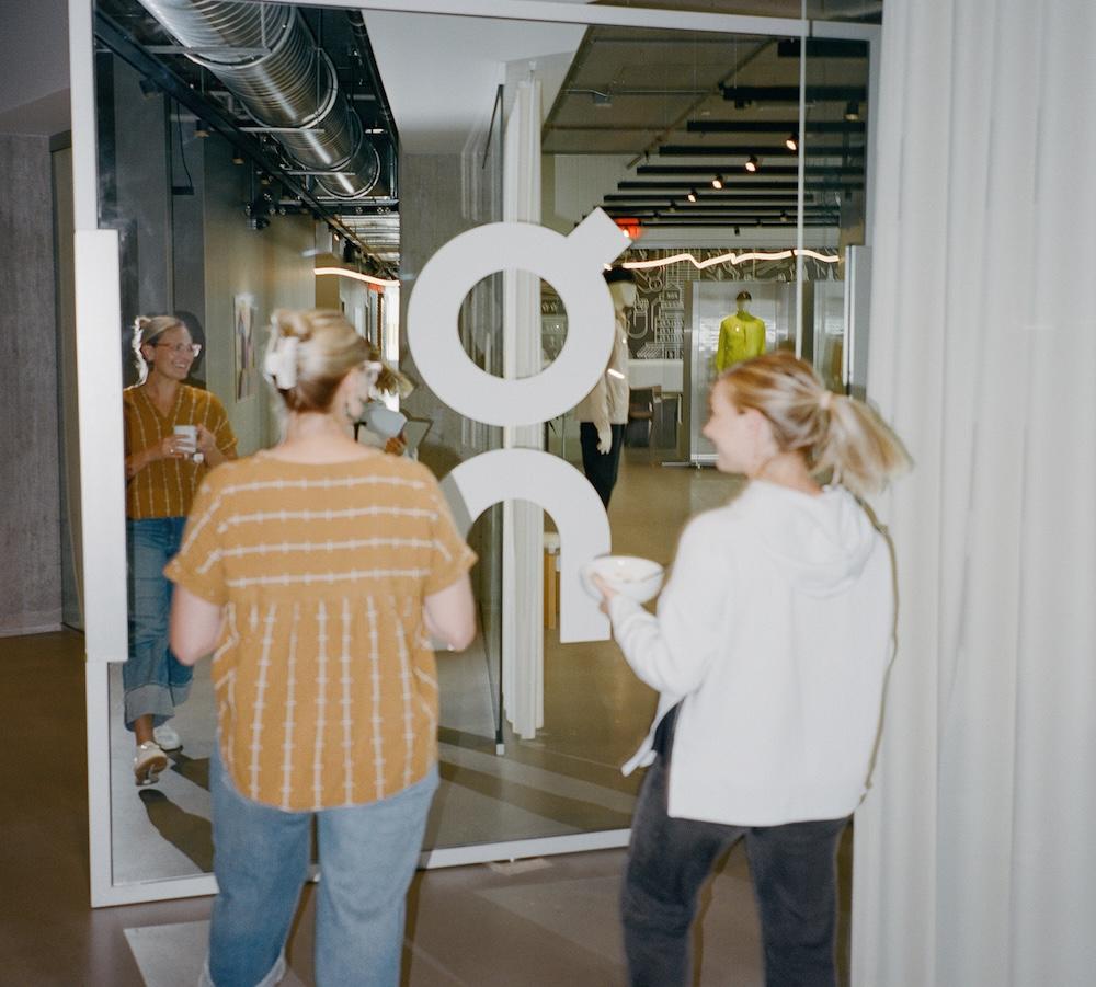 Two people enter through a glass door bearing a large white circular logo, one holding a bowl, a mirrored reflection of another person is visible, and a mannequin wearing bright yellow stands in the background.