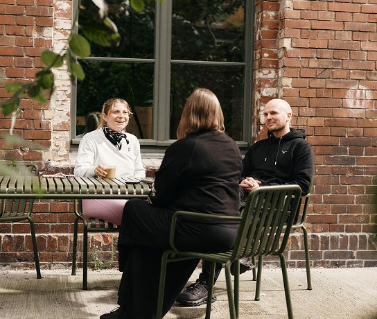 Three people sit around a green metal outdoor table in front of a brick building, one holding a takeaway cup.