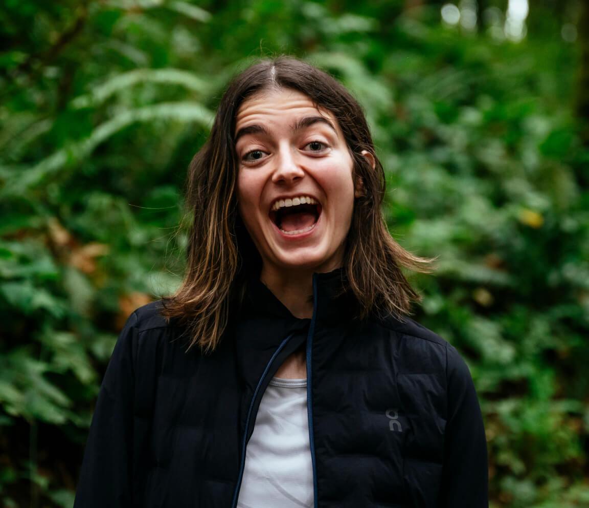 Young woman with shoulder-length hair smiling widely with her mouth open, standing in front of dense green foliage while wearing a dark jacket.