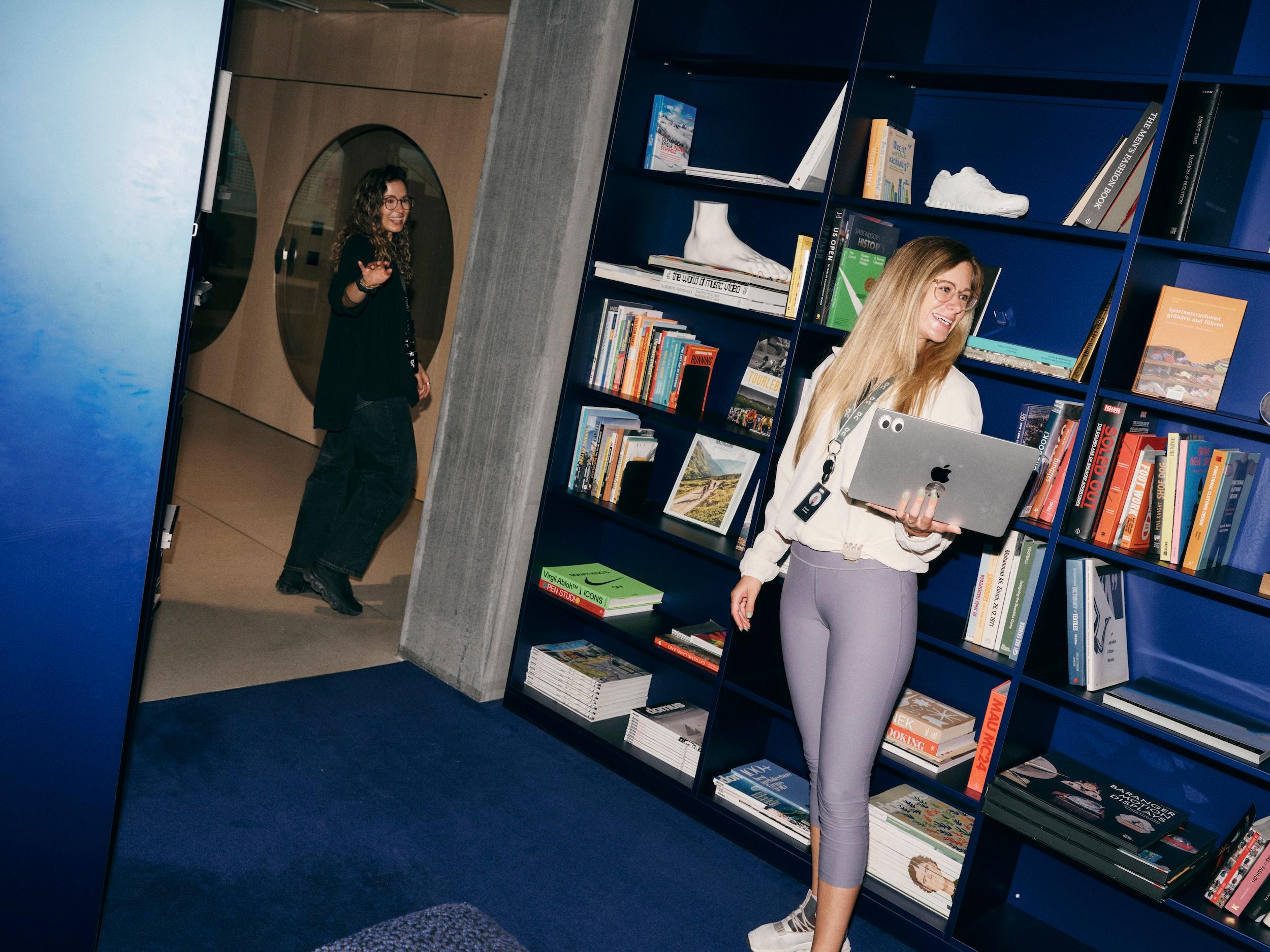 Two women in a room with blue bookshelves, one smiling and holding a laptop by the shelves while the other waves as she walks through a doorway.