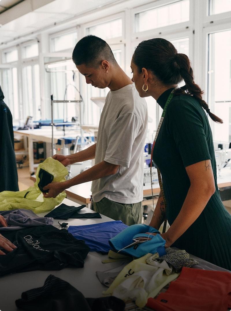 Two designers examine fabric samples in a sunlit studio with sewing machines.