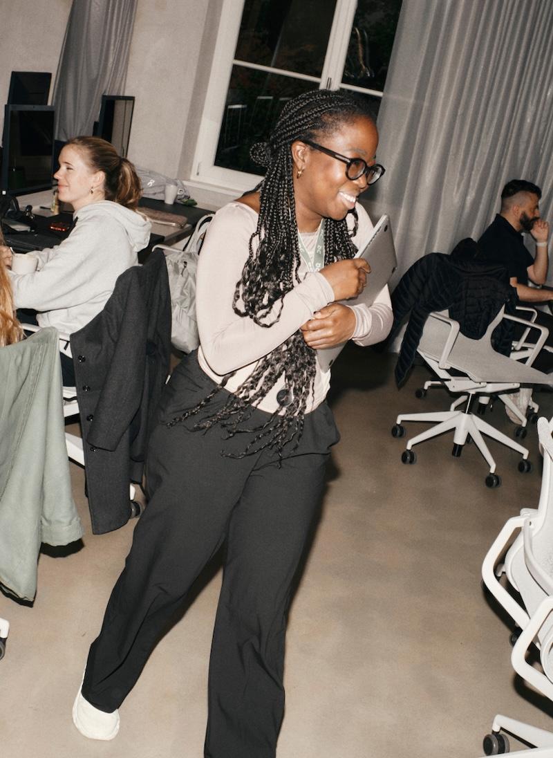 Smiling woman walks through an office holding a closed laptop, surrounded by coworkers at desks.