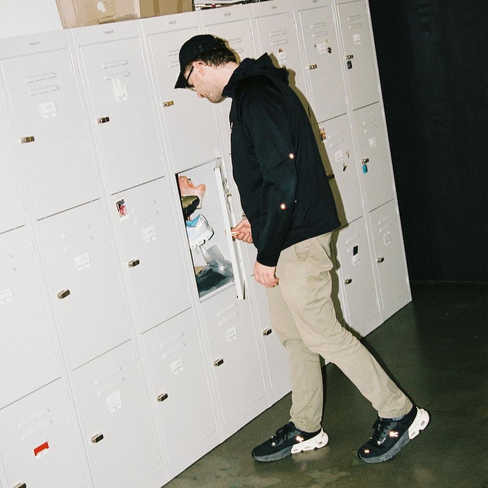 A man wearing a black cap, black hoodie and khaki pants opens and looks into a small white locker in a row of white lockers.
