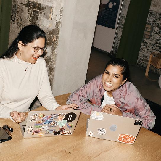 Two people sit at a wooden table smiling while using sticker-covered laptops in an indoor workspace with exposed brick walls.