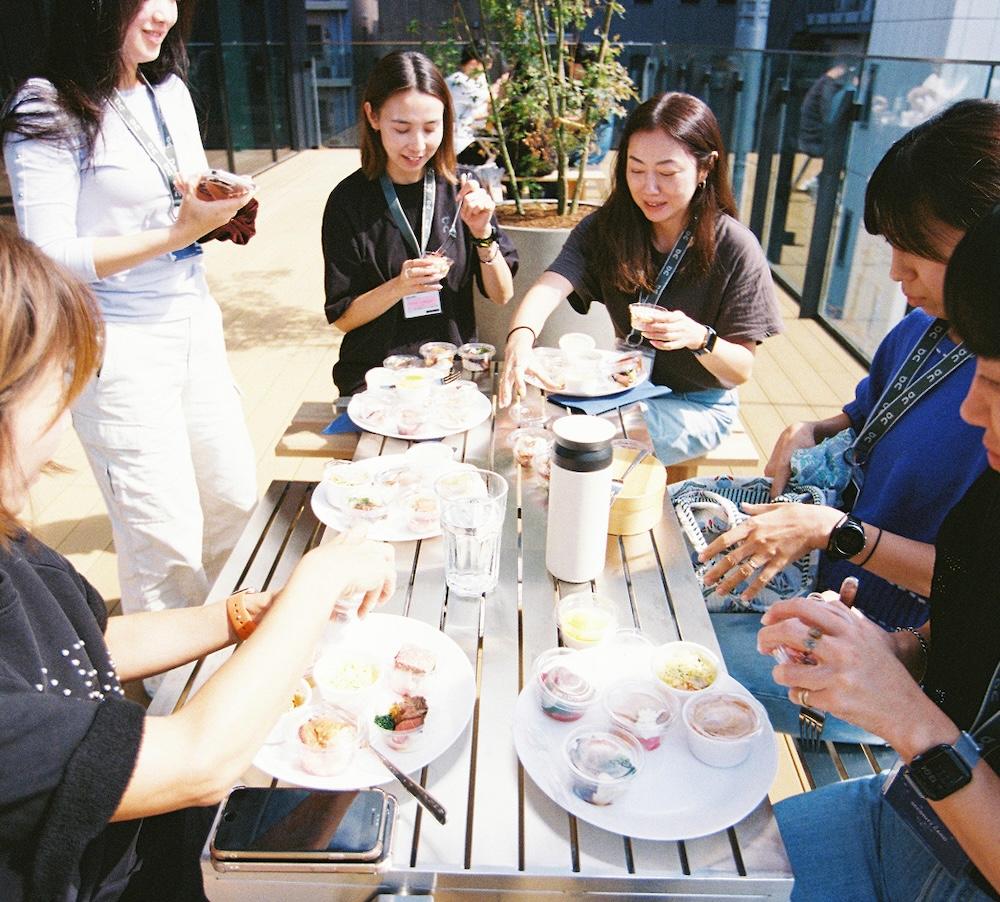 A group of people wearing lanyards sit and stand around an outdoor table, eating from plates and small cups of food.