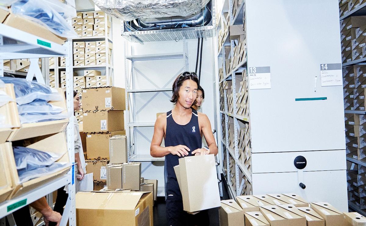Two people stand in a storage room lined with shelves and stacks of cardboard boxes, with the person in front holding a box.