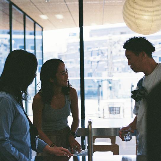 Three people talk in a sunlit room by a large window, with one person holding a glass of water.