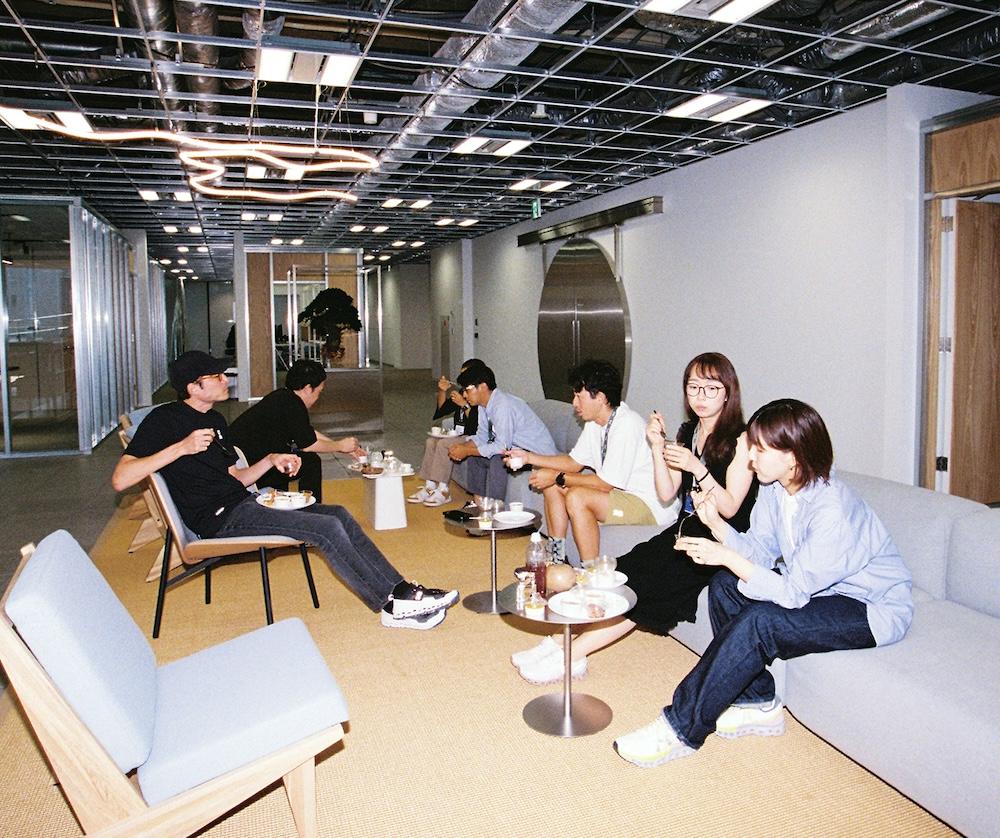 Several people sit on sofas and chairs in a modern open office lounge with an exposed ceiling, eating and talking around small round tables.