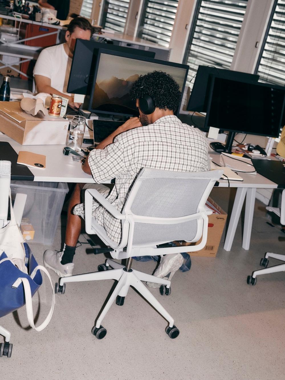 Person wearing headphones works at a computer in a busy office with desks, packaging boxes, and supplies.