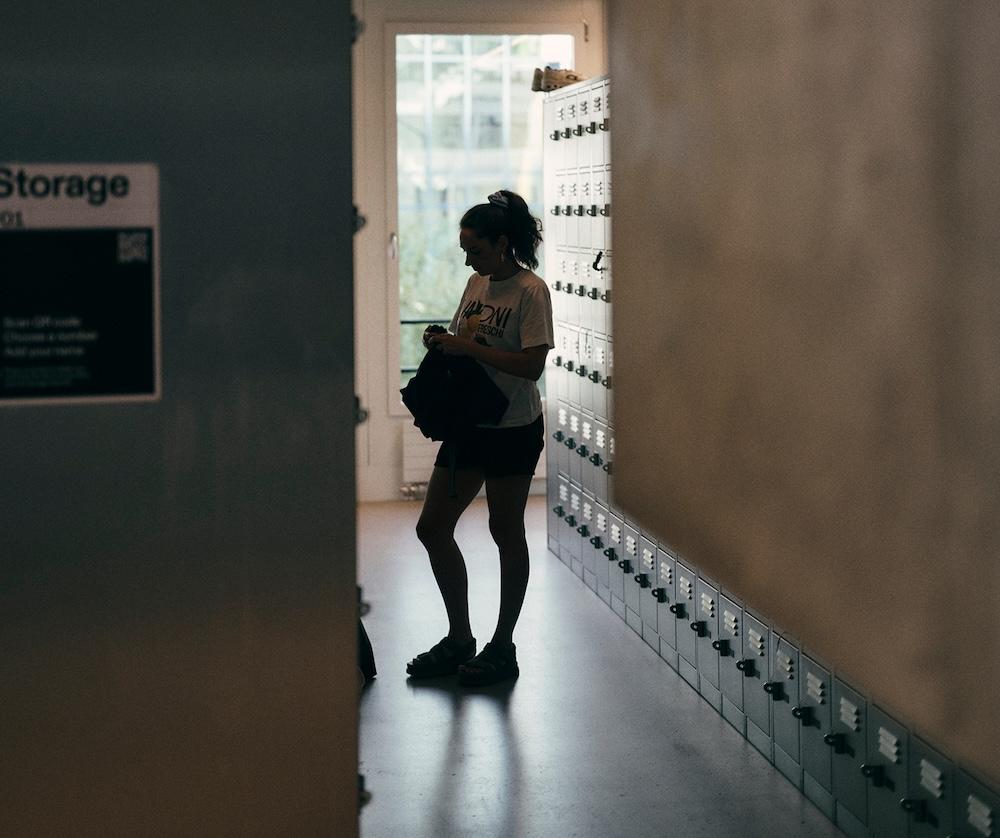 A person in a T-shirt and shorts stands in a dim hallway next to a row of lockers holding a bag and looking down.
