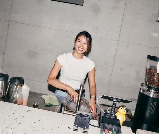 A smiling woman stands behind a countertop washing a blender pitcher at a sink with coffee equipment and a small yellow rubber duck on the counter.
