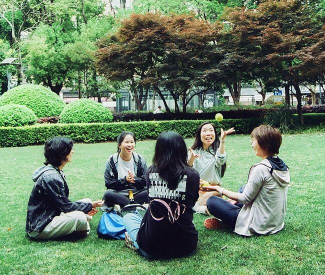 Five people sit in a circle on the grass of a city park, smiling and talking beneath trees with a high-rise building in the background.