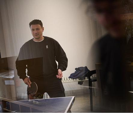 A person in a dark sweatshirt holds a table tennis paddle while standing beside an indoor table tennis table.