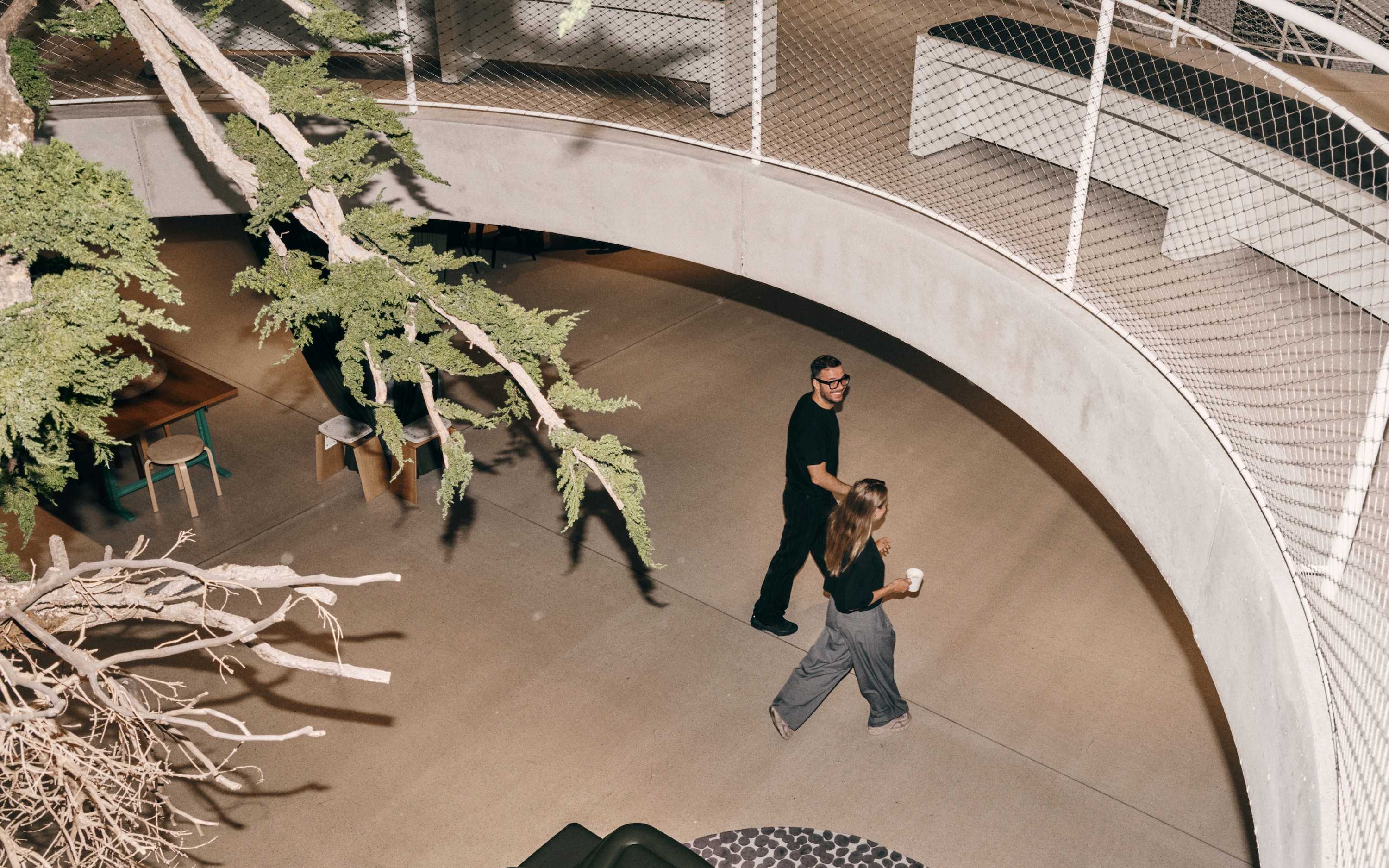 Two people walk beneath a curved concrete balcony in a modern, indoor space with trees and lounge furniture.