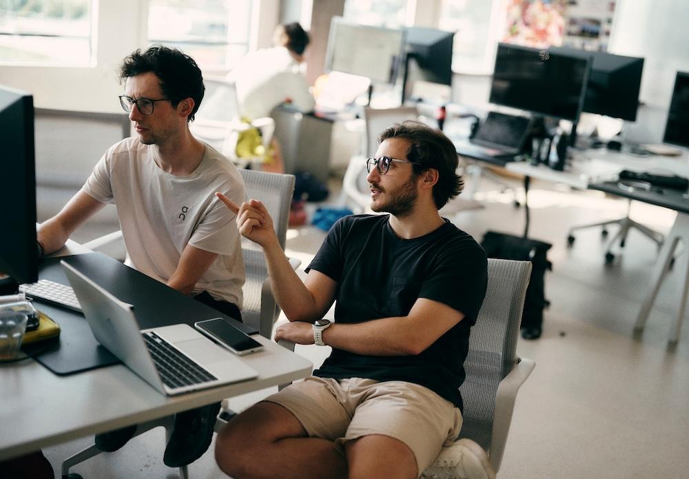 Two men wearing glasses sit at a desk in an open office, with one man pointing toward a computer screen.