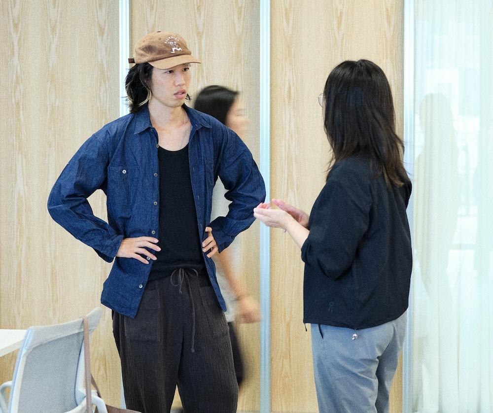 A man in a brown cap and blue button-up shirt stands with his hands on his hips facing a woman wearing glasses and a black jacket who is gesturing with her hands in an indoor room.