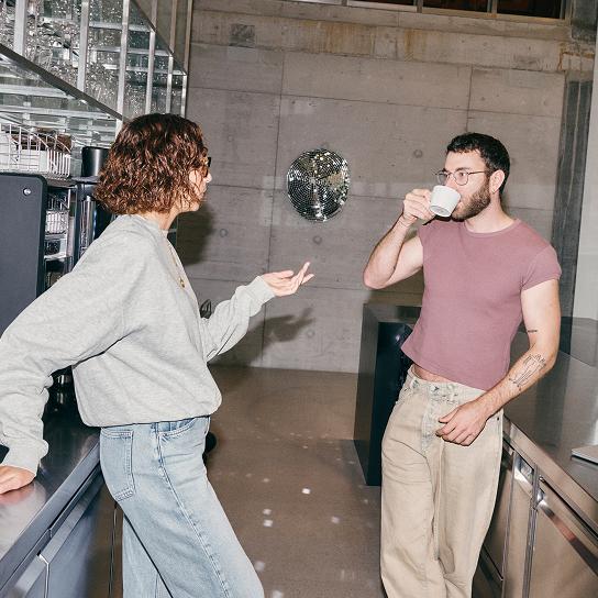 A woman in a gray sweatshirt and jeans leans on a counter while talking to a bearded man in glasses and a pink T‑shirt who sips from a white mug in a room with a concrete wall and a small disco ball.