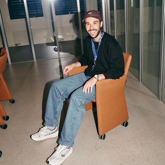 A smiling man wearing a cap, lanyard, black sweatshirt, jeans and white sneakers sits in an orange rolling chair in a glass-walled indoor space.