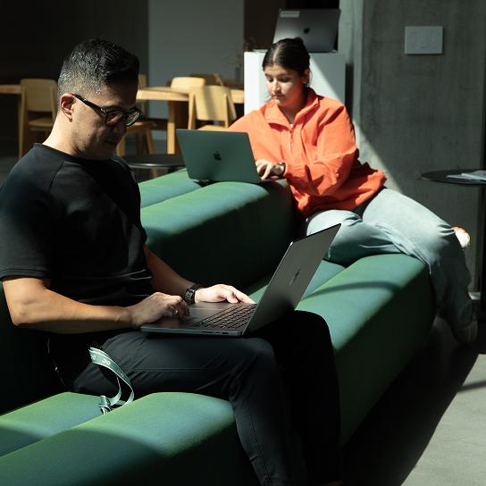 Two people sit on a green modular sofa using laptops, one wearing a black shirt and glasses and the other wearing an orange jacket.