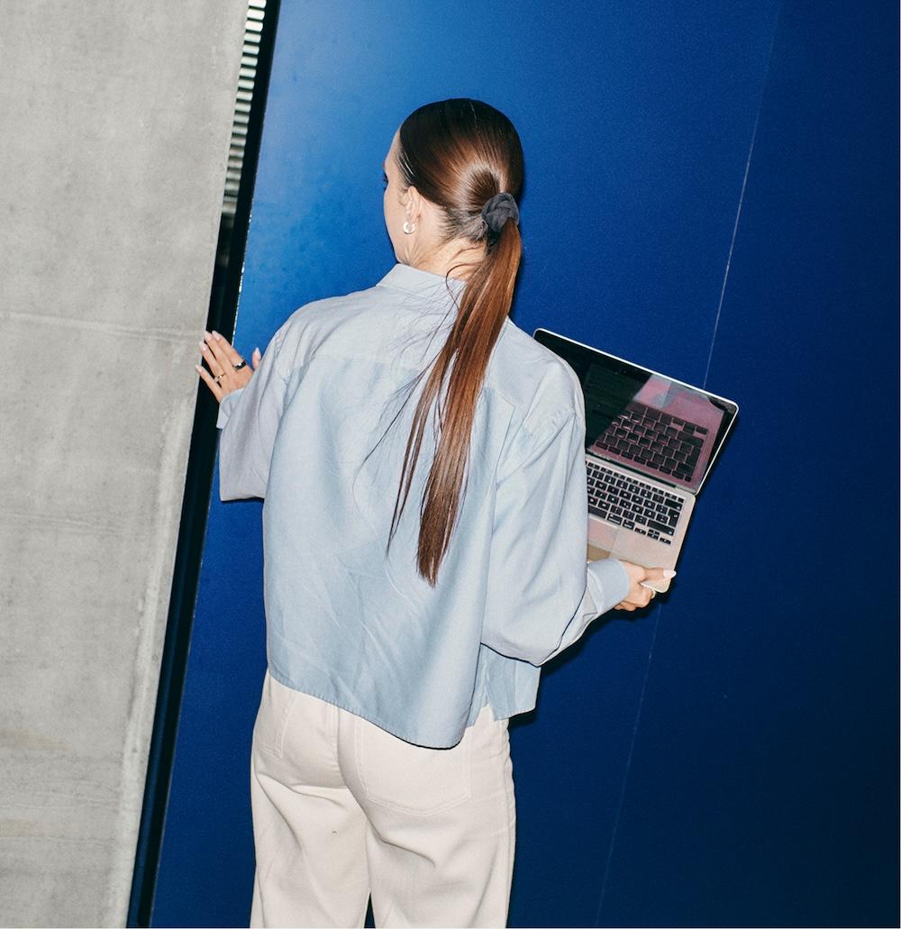 Person seen from behind with long ponytailed hair, wearing a light blue shirt and white pants, holding an open laptop against a blue wall.