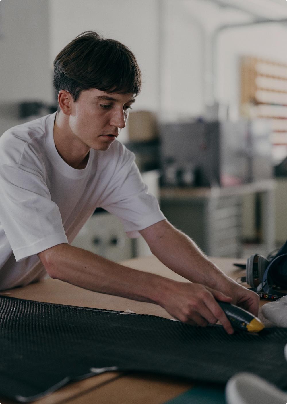 A young man in a white T-shirt leans over a table and uses a handheld utility knife to cut a piece of black textured material.