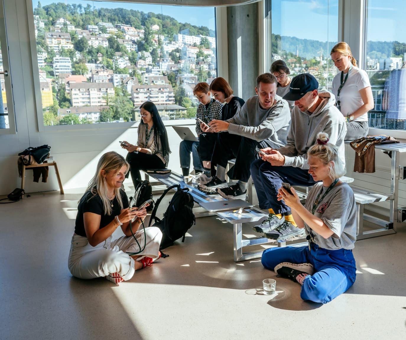A group of people sat chatting whilst on their devices in a room with large windows that's flooded with light.