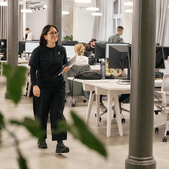 A woman in glasses and black clothes walks through a modern open-plan office holding a laptop while coworkers sit at desks with computer monitors.