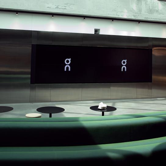 Modern interior with curved green bench seating, round black tables, a large black wall-mounted screen showing two identical white stylized symbols, and a small white object on one table.
