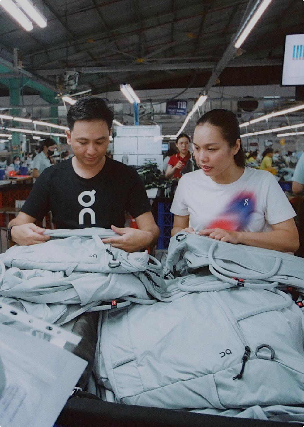 Two people inspect light gray backpacks on a table inside a busy manufacturing facility.
