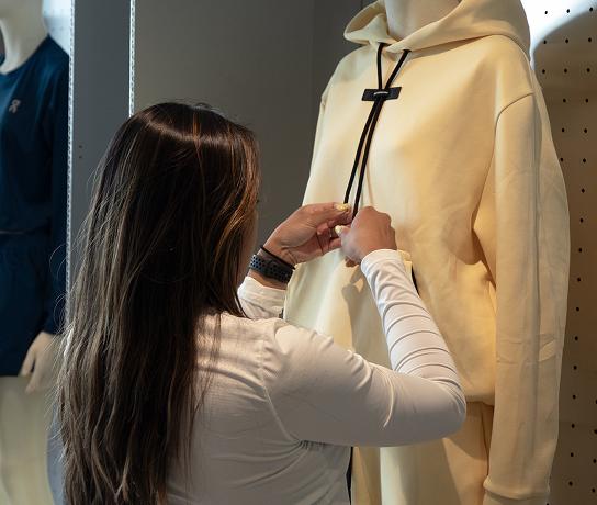 A person adjusts the black drawstrings of a pale yellow hoodie on a mannequin in a clothing store.
