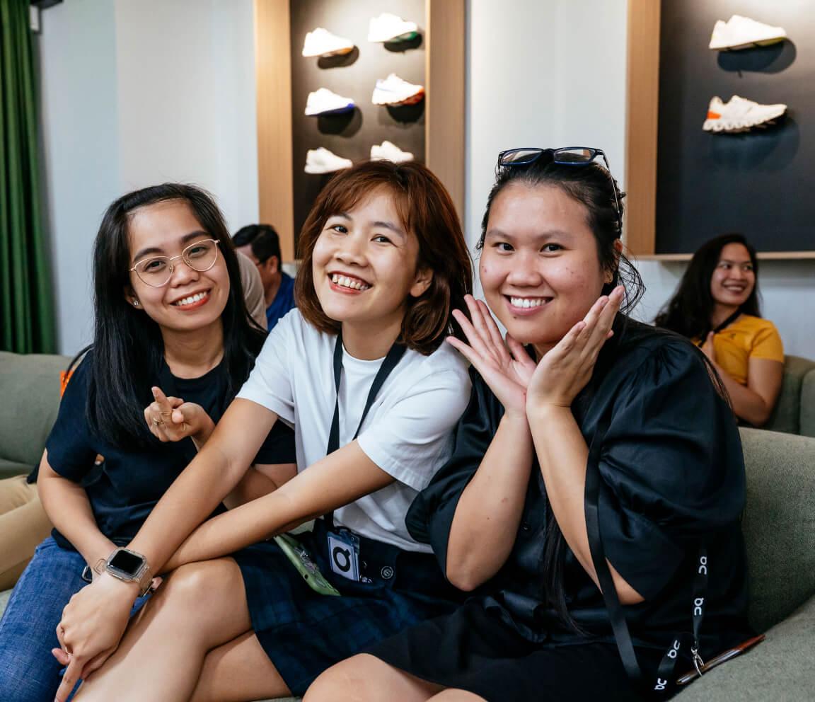 Three women smiling and posing on a couch in a modern room with sneaker displays on the wall behind them; one woman makes a heart gesture and another rests her face in her hands.